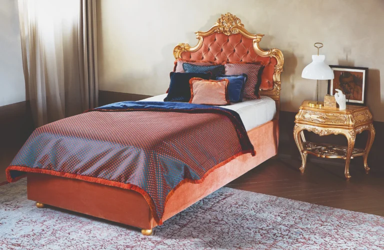 A single baroque-style bed with a terracotta velvet base, a gilded carved headboard, and a burgundy-blue checkered bedspread.