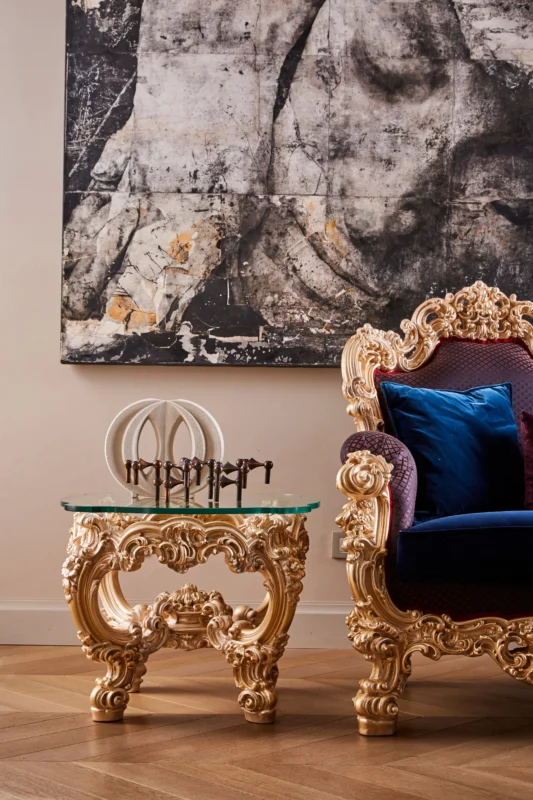 Detail: a gilded baroque coffee table with a glass tabletop next to a blue velvet armchair, with decorative items placed on the table.