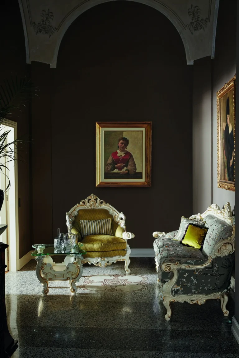 A dark living room with two baroque armchairs - one in olive velvet and one in gray-blue with a floral print - and a glass coffee table on a white carved base.