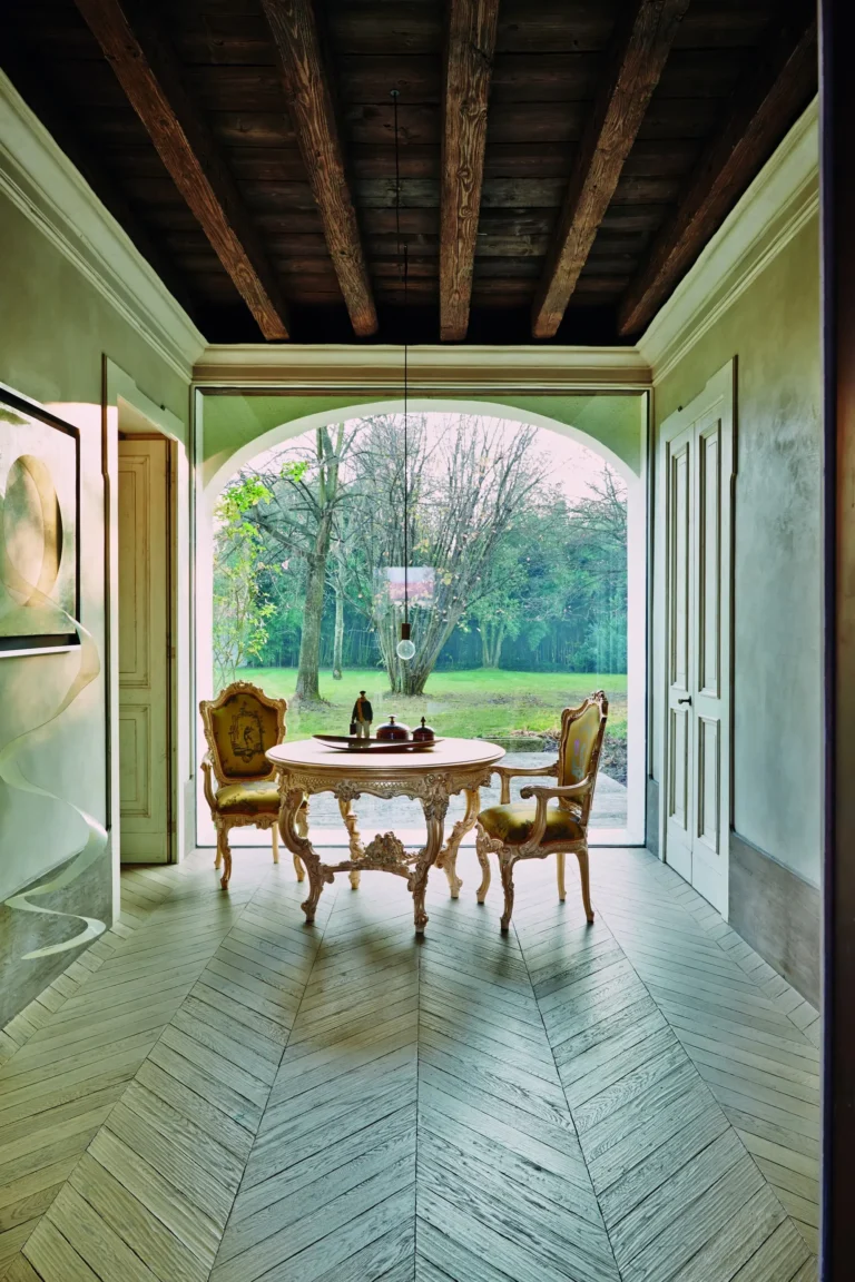 Elegant interior with a baroque round table and two olive-upholstered armchairs by a floor-to-ceiling arched window overlooking the park, with wooden ceiling beams.