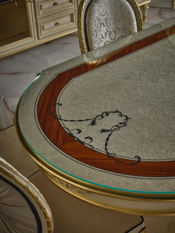 Close-up of an oval dining table with an inlaid top — frosted glass, a mahogany strip, and a blue floral ornament framed by a gilded border.