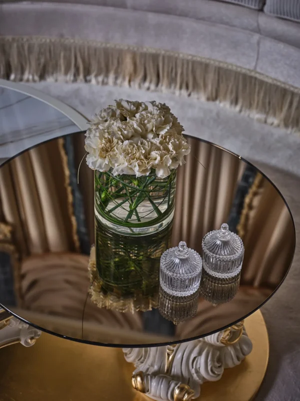 Round coffee table with a bronze mirrored tabletop, a glass vase with white carnations, and two crystal jars with lids.