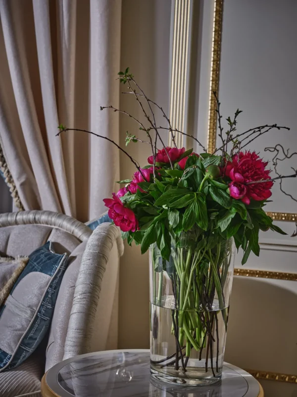 Close-up of a glass vase with a lush bouquet of crimson peonies and branches, set against beige curtains and a gilded mirror.